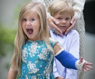 Girl in floral dress with blue cast, mouth open in excitement; boy behind her looking shy, hands on his face.
