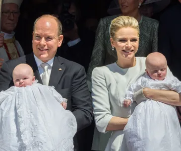A man and woman holding two babies in white baptismal gowns, smiling outdoors.