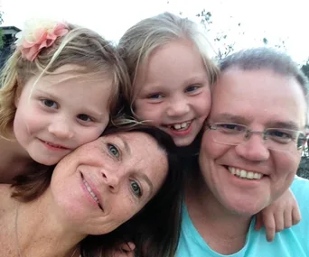 Family selfie with a man, woman, and two smiling young girls, one with a flower in her hair, all closely huddled.