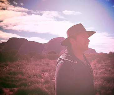 A man in a wide-brimmed hat stands in an outback landscape with rock formations in the background under a cloudy sky.