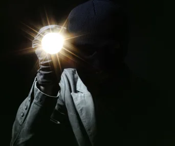 A person in a ski mask holding a bright flashlight in a dark setting.