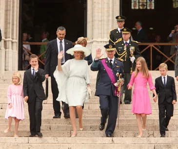 Royal family descending stairs on Belgium's National Day; King, Queen, and children in formal attire, greeting the crowd.
