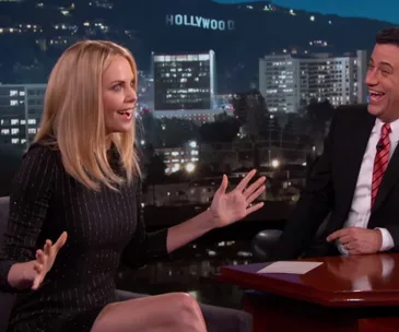 Charlize Theron animatedly talks on a late-night show, cityscape in background.