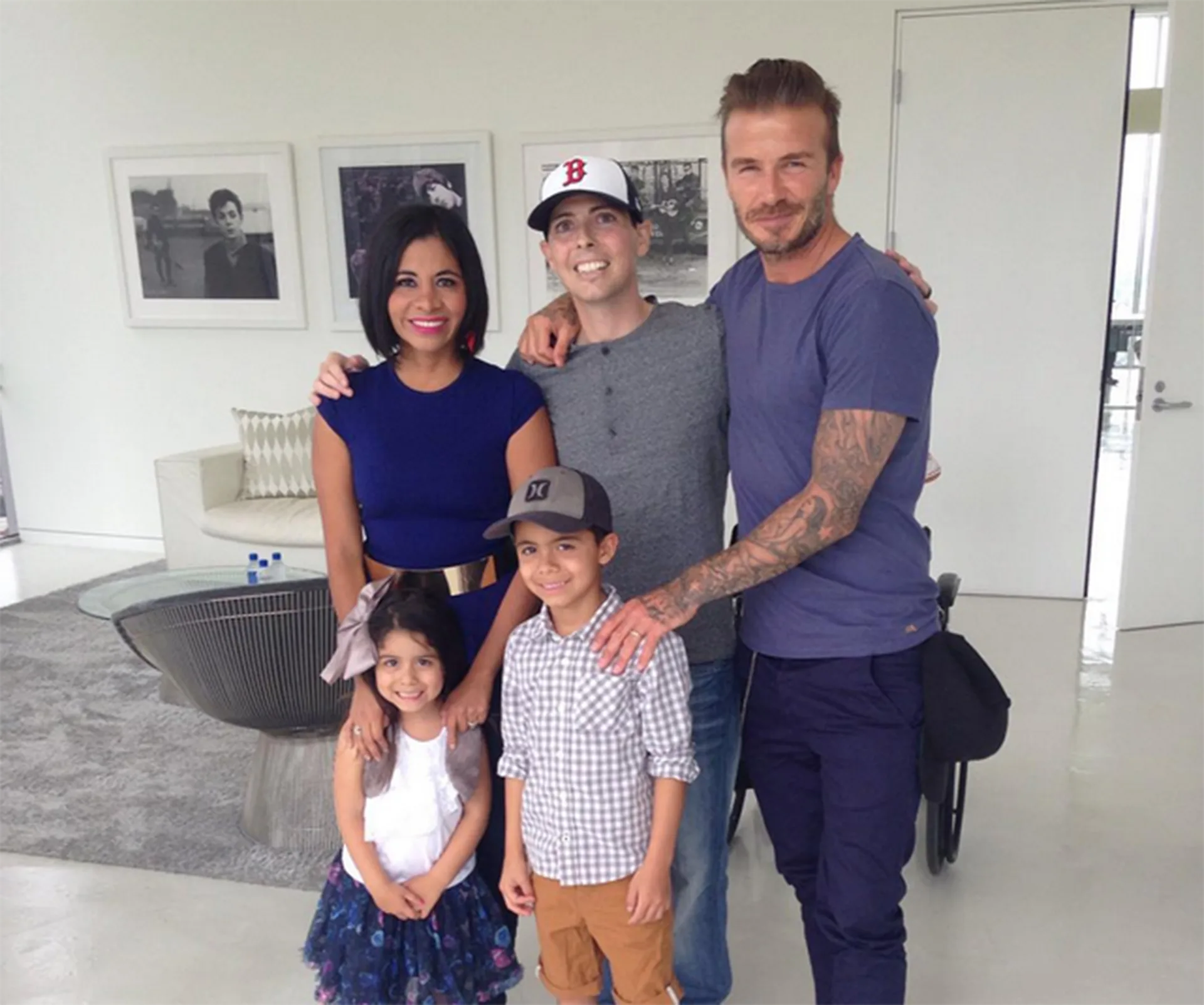 Family of six, including David Beckham, smiling together indoors with framed photos on the wall in the background.