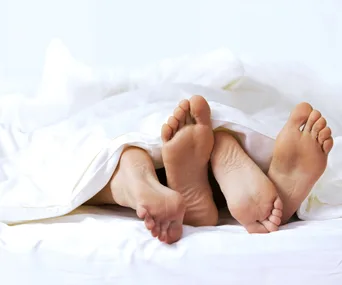 Two pairs of feet sticking out from under a white bedsheet, suggesting intimacy and relaxation.
