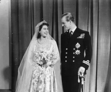 A bride in an elegant gown stands beside a groom in a naval uniform, both smiling, in a historic wedding photograph.