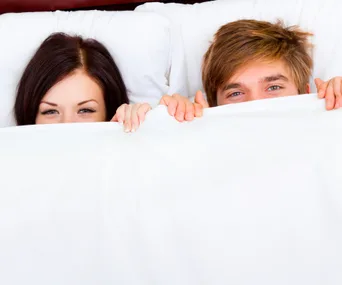 Couple smiling under white bed sheets, eyes visible and peeking.