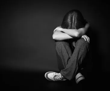 A person sitting on the floor, hugging their knees with their head down, in a dark and somber setting.