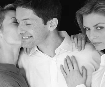 Three people in black and white, one kissing a smiling man's cheek, another looking serious on the right.