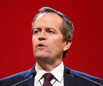 A person speaking at a public event against a red background, wearing a suit and tie.