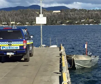 Police vehicle on a pier next to a small boat on the water with hills and houses in the background.