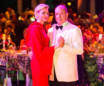 A couple dancing at a formal event; woman in red jumpsuit, man in white tuxedo with bow tie, surrounded by seated guests.