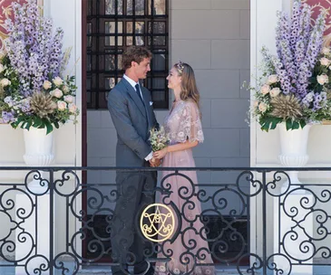 A couple stands on a decorated balcony, surrounded by flower arrangements, lovingly gazing at each other.