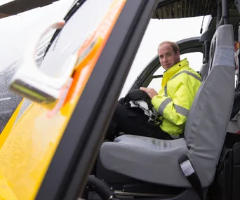A person in a yellow jacket sits in a helicopter cockpit holding a helmet, with raindrops visible on the window.