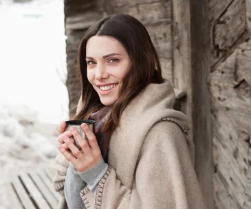 Woman wrapped in a beige blanket, smiling, and holding a cup outdoors by a wooden wall in winter.