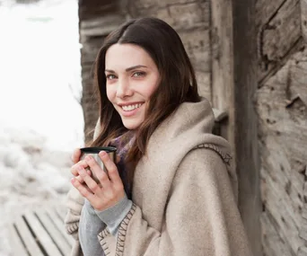 Woman wrapped in a beige blanket, smiling, and holding a cup outdoors by a wooden wall in winter.
