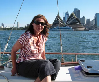Woman in sunglasses on a boat, with the Sydney Opera House and city skyline in the background on a sunny day.