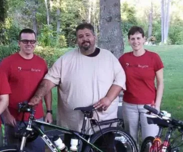 Man standing with two people and a bicycle, outdoors, surrounded by trees.