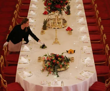 A staff member sets an ornate banquet table with red chairs and floral centerpieces in Buckingham Palace.