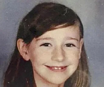 A smiling young girl with long hair, wearing a gray top against a plain background.