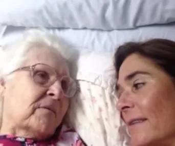 Elderly woman with glasses in bed, looking at middle-aged woman lying beside her, both appear to be engaged in conversation.