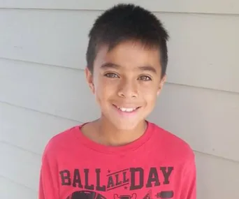 A smiling young boy in a red "BALL ALL DAY" shirt stands against a light-colored wall.