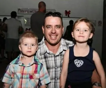 A man smiling with two young children, one on each side, in a casual indoor setting.