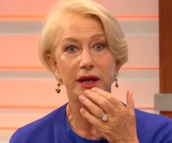 A surprised woman in blue on a TV set, holding a hand to her mouth.