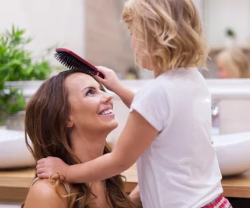 A woman is kneeling, smiling at a young girl who is brushing her hair in a bright bathroom setting.
