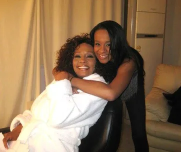 Two women, one in a white robe, are hugging and smiling warmly at the camera in a cozy room.