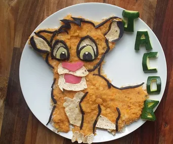 Food art depicting Simba from "The Lion King" made with mashed potatoes and vegetables spelling "Jacob" on a plate.