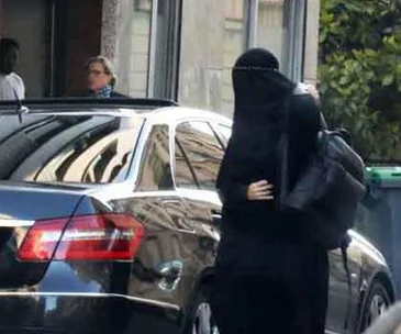 A person in a burqa stands near a black car on a city street.