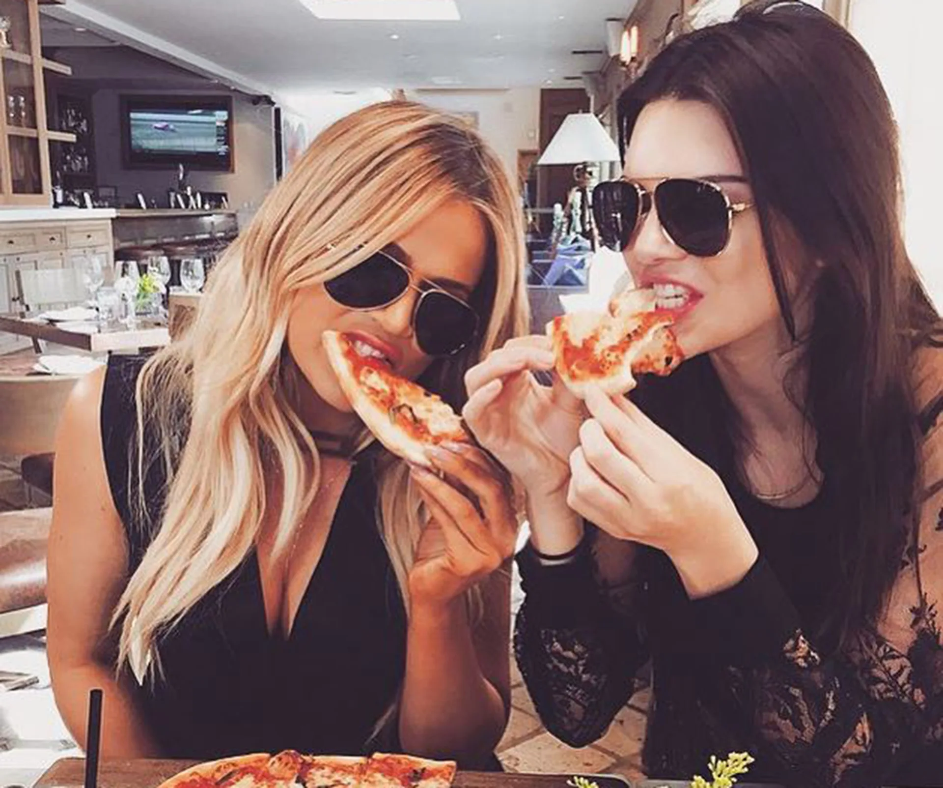 Two women wearing sunglasses enjoying slices of pizza at a restaurant.