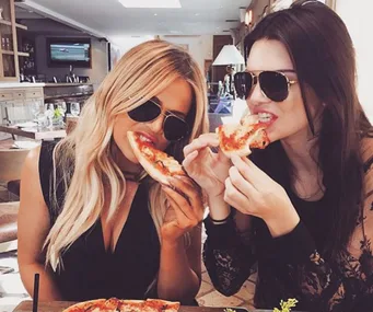 Two women wearing sunglasses enjoying slices of pizza at a restaurant.