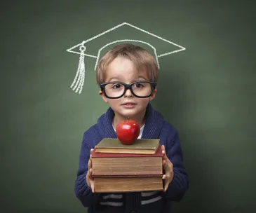 A young child with glasses holds books, topped with an apple, in front of a chalkboard with a drawn graduation cap.