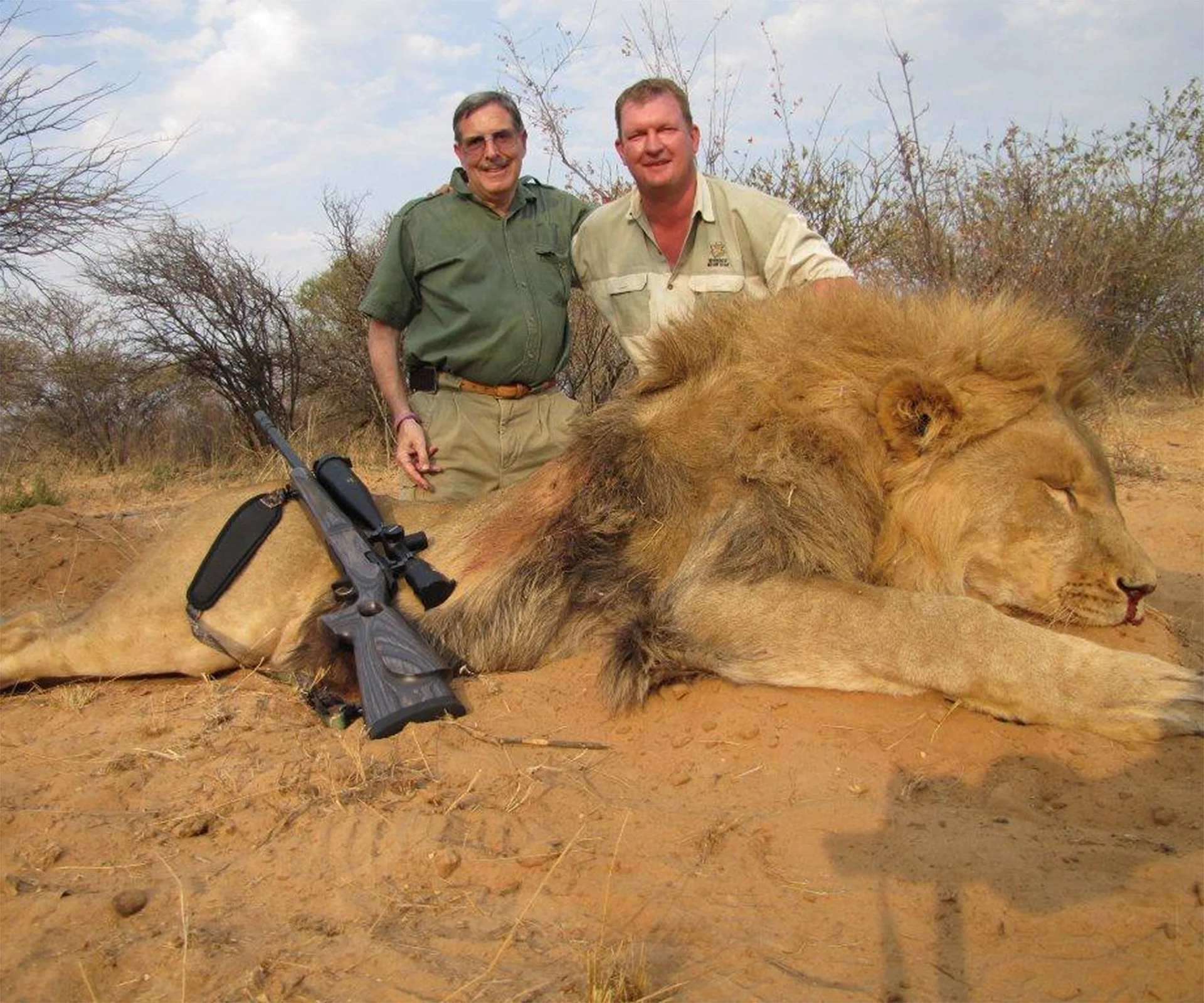 Two men pose next to a lion lying on the ground, with a rifle resting on the lion's body.