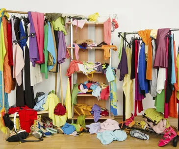 Cluttered room with colorful clothes, shoes, and accessories scattered around a wardrobe and rack.