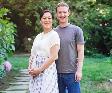 A couple poses in a garden; the woman, wearing a dress, is visibly pregnant, and they stand on a stone path surrounded by greenery.