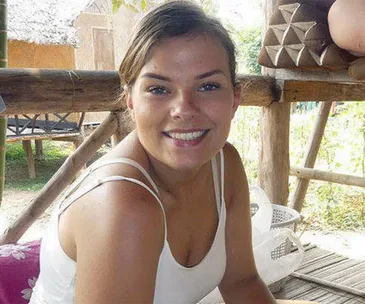A woman smiling, sitting on a wooden structure outdoors in a casual setting.