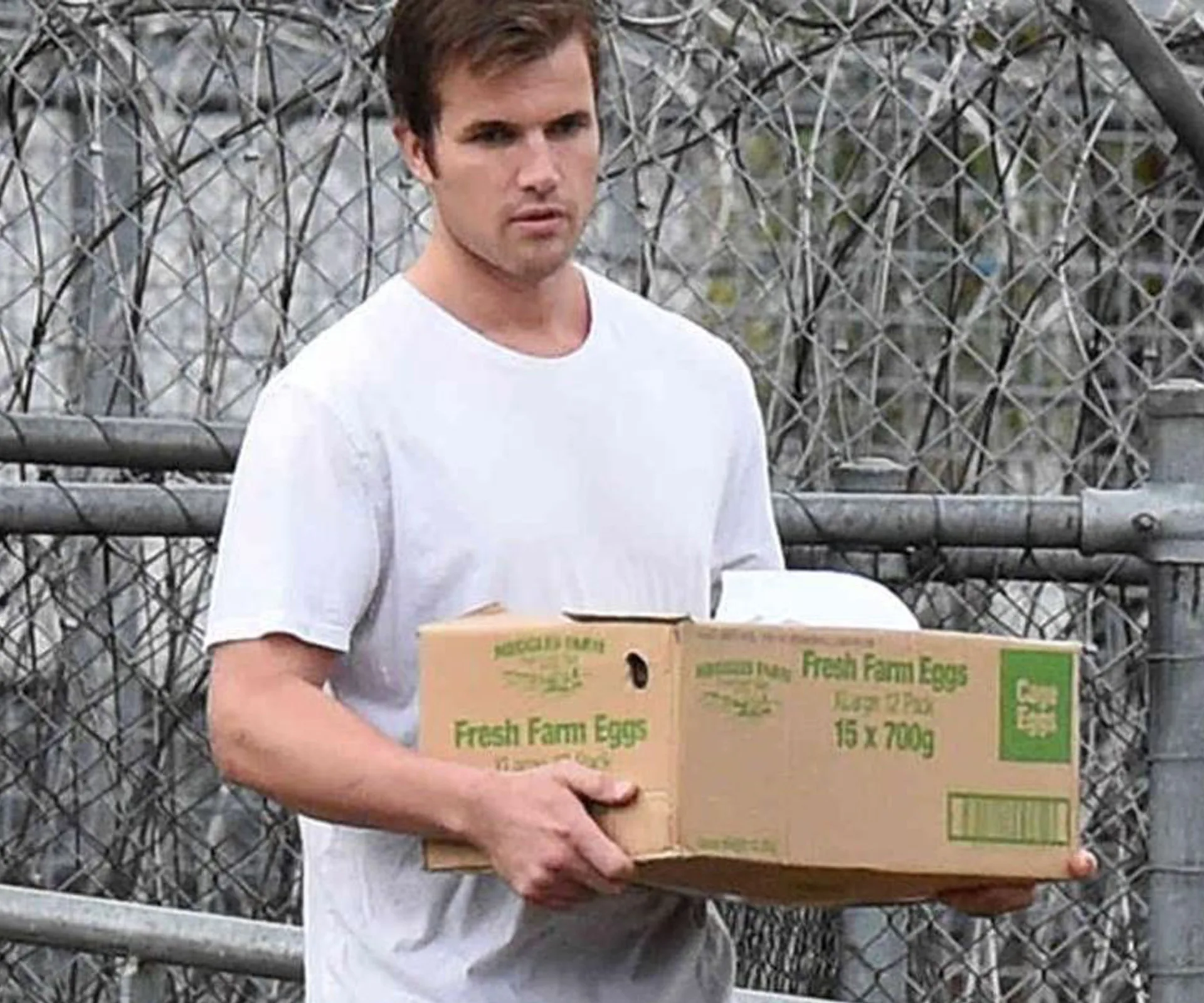 Man in white shirt carrying a box of eggs near a chain-link fence.