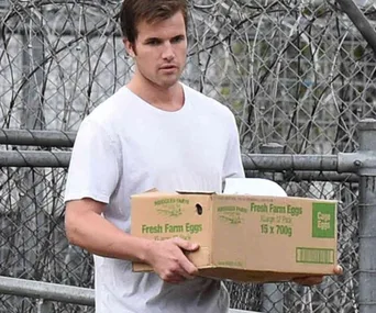 Man in white shirt carrying a box of eggs near a chain-link fence.