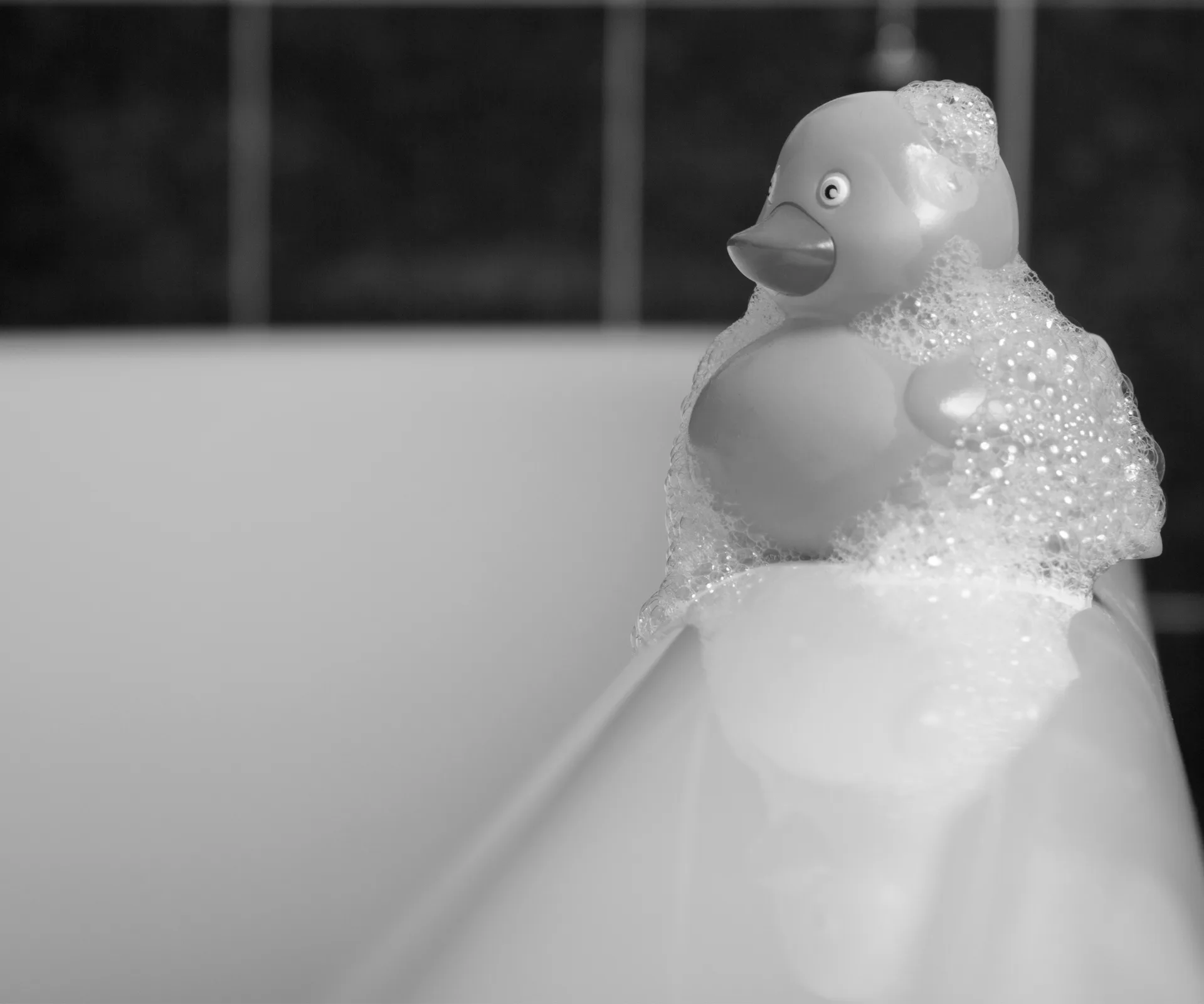 Rubber duck covered in bubbles, resting on the edge of a bathtub.