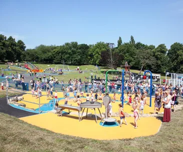 Children playing in a sunny park playground with swings and splash area, surrounded by adults and trees in the background.