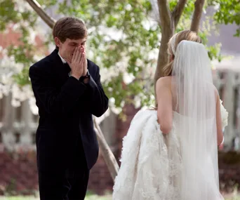 A groom in a tuxedo, hands on face, and a bride in a wedding dress with veil, outdoors.