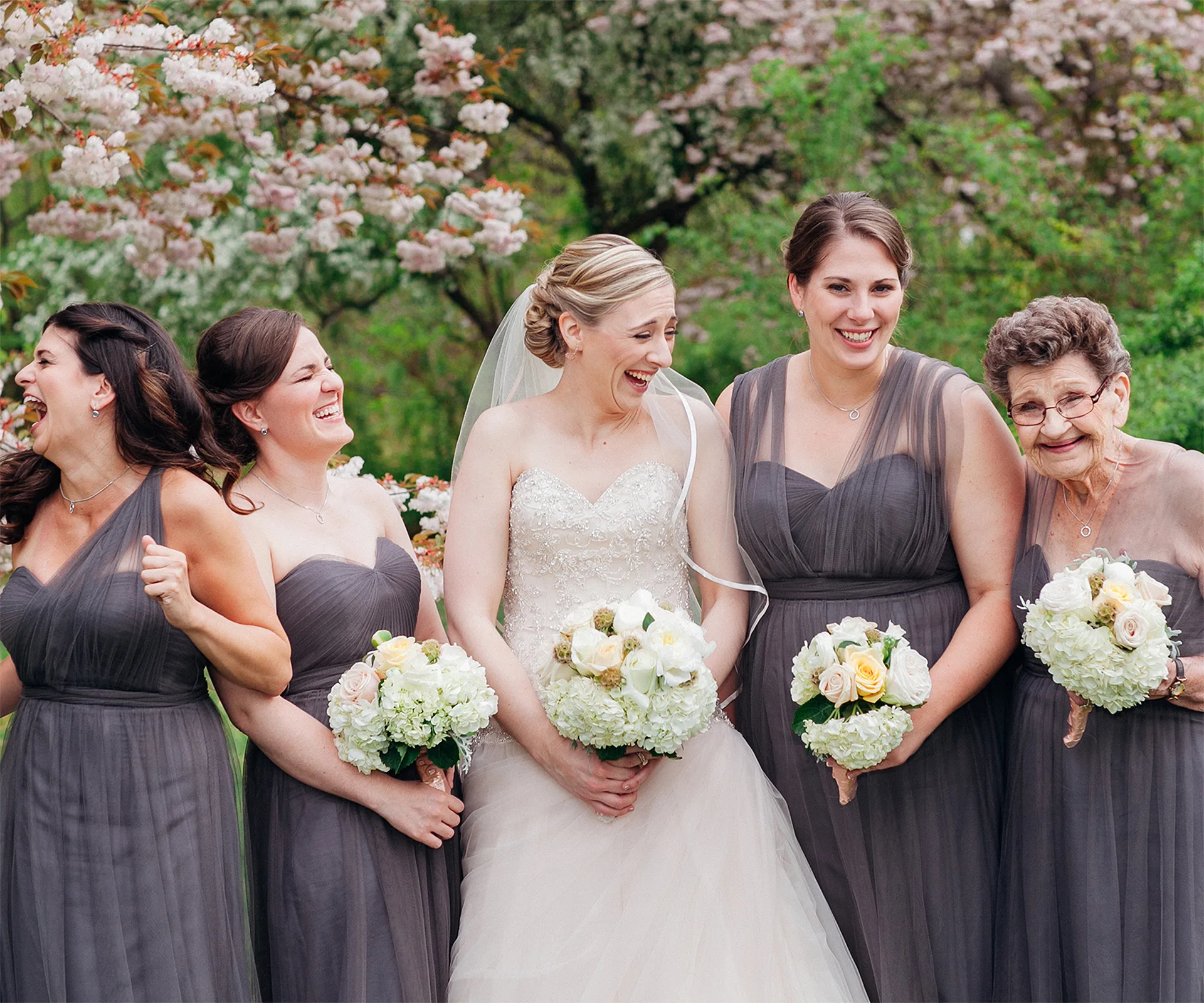 Bride and bridesmaids in gray dresses laugh together, holding bouquets, with blooming trees in the background.