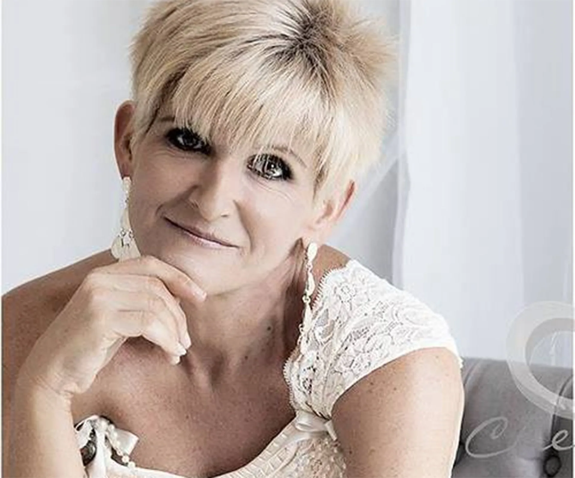 A smiling woman with short blonde hair and lace dress, seated indoors with hand near face.