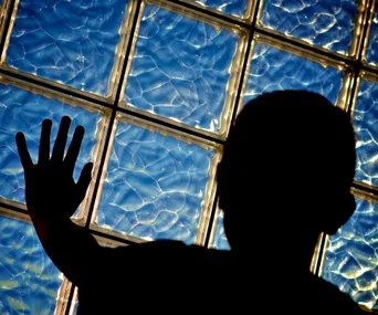 Silhouette of a child with hand against a glass block wall, under blue, wavy light.