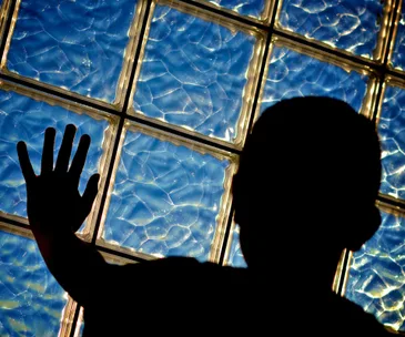 Silhouette of a child with hand against a glass block wall, under blue, wavy light.