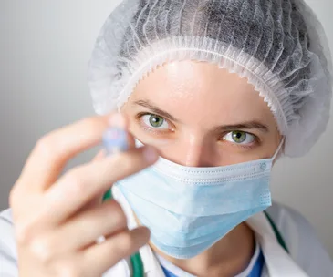 Nurse wearing a face mask and hairnet, focusing on holding a small vial or syringe.