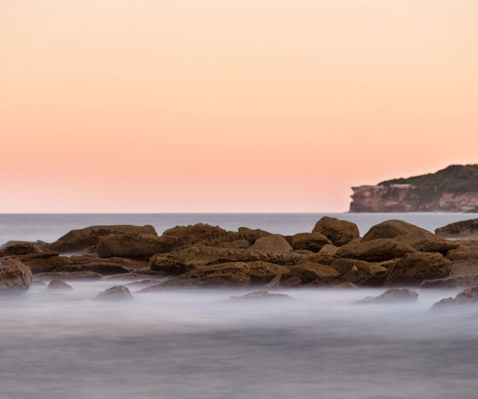 Rocky beach at sunset with calm waves and a distant cliffside under a pastel sky.